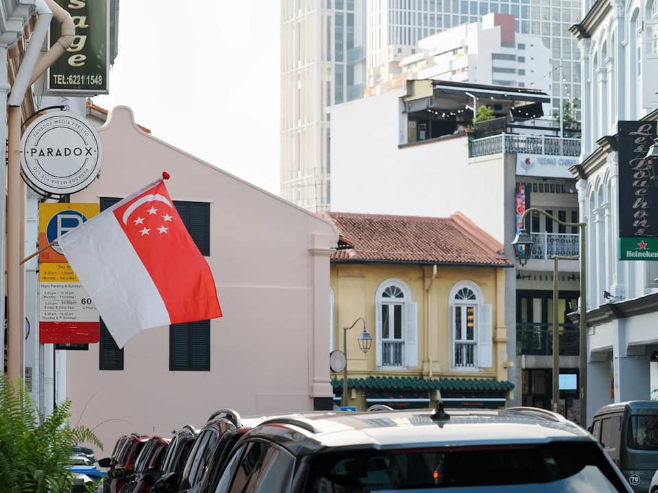 The Singapore flag flying on a street with historic shophouses and parked cars