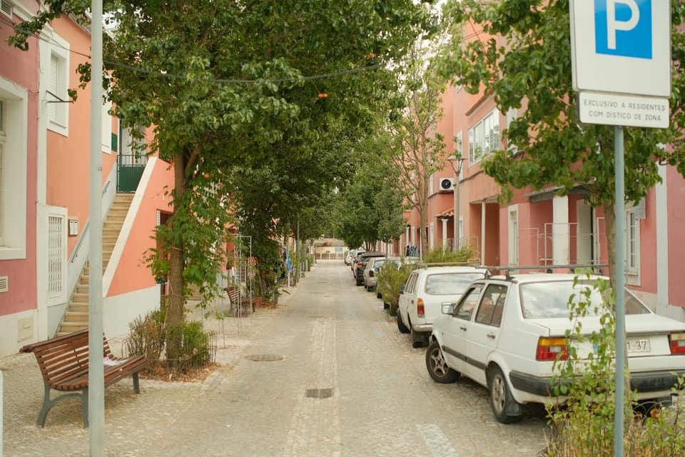 A narrow cobblestone street lined with pink buildings and parked cars under green trees